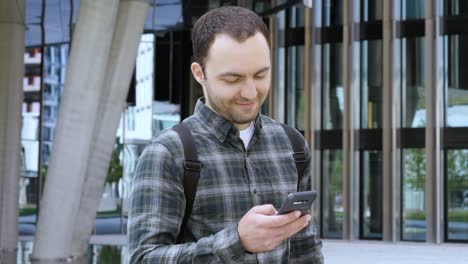 Hombre-Casual-Caminando-cerca-del-Edificio-Moderno-y-usando-smarphone-sonriendo