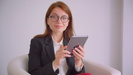 Retrato-de-primer-plano-de-joven-empresaria-caucásica-en-gafas-enviando-mensajes-de-texto-en-la-tableta-mirando-ta-cámara-sonriendo-felizmente-sentado-en-el-sillón