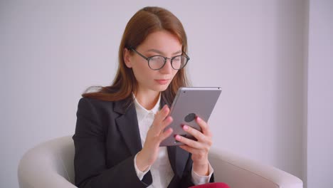 Retrato-de-primer-plano-de-joven-alegre-empresaria-en-gafas-escribiendo-en-la-tableta-mirando-ta-cámara-sonriendo-felizmente-sentado-en-el-sillón