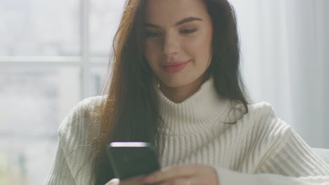 Schöne-junge-Frau-mit-Smartphone-lächelnd,-während-auf-dem-Stuhl-sitzen.-Sinnliche-Mädchen-tragen-Pullover,-Surfs-Internet,-Beiträge-auf-Social-Media-beim-Entspannen-in-cozy-Apartment.-Closeup-Portrait