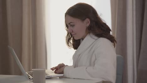 Close-up-of-positive-Caucasian-girl-using-laptop-in-the-morning.-Young-beautiful-woman-in-white-bathrobe-using-social-media.-Internet,-online,-lifestyle,-leisure.