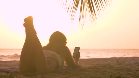 CLOSE-UP:-Young-woman-lies-on-the-beach-and-surfs-the-internet-on-smartphone.