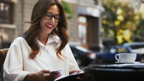 Geschäftsfrau-sitzt-am-Tisch-mit-tasse-Kaffee-im-Straßencafé.-Details-des-Startups-auf-Dem-Papier-analysieren,-aufs-Smartphone-schauen,-jubeln.-Nahaufnahme