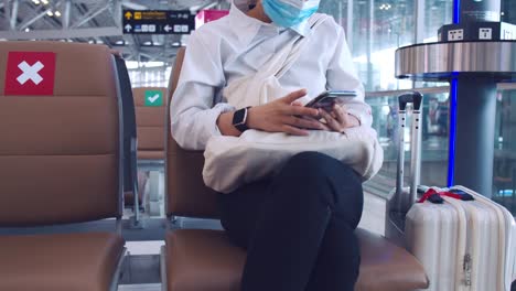 Businesswoman-wearing-surgical-mask-protection-and-use-smartphone-checking-social-media-feeds-while-sitting-a-chair-in-the-airport-terminal-and-keep-a-social-distance-during-the-COVID-19-pandemic.