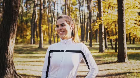 Young-woman-smiling-and-listening-to-music-through-earphones-while-posing-in-autumn-forest-on-sunny-day