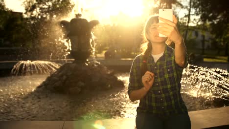 A-young-woman-takes-a-selfie-on-a-phone-near-a-fountain-in-a-public-park