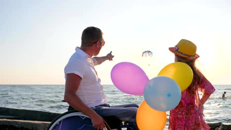 disabled-man-pointing-finger-in-blue-sky-on-color-parachute,-father-in-wheelchair-with-child-near-sea