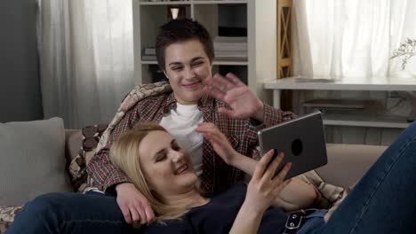 Lesbian-couple-is-resting-on-the-couch,-and-having-video-conversation-with-friend,-waving-their-arms,-video-call-60-fps