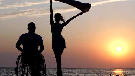 shapely-girl-is-standing-on-jetty-and-waving-cloth-in-front-of-man-disabled-into-wheel-chair-on-background-of-sunset-at-summer-weekend