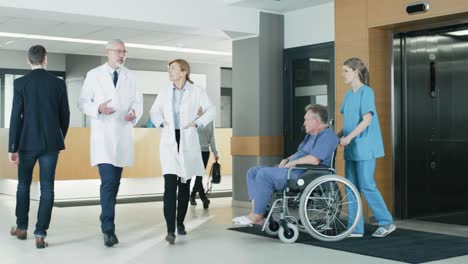 In-the-Hospital,-Doctors,-Nurses-and-Personnel-Busy-Working,-Receptionist-Talks-with-a-Patient,-Assistant-Moves-Elderly-Man-in-the-Wheelchair.-New-Modern-Medical-Hospital-with-Professional-Staff.