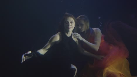 Two-attractive-woman-in-chiffon-dress-swimming-together-underwater-in-dark-pool