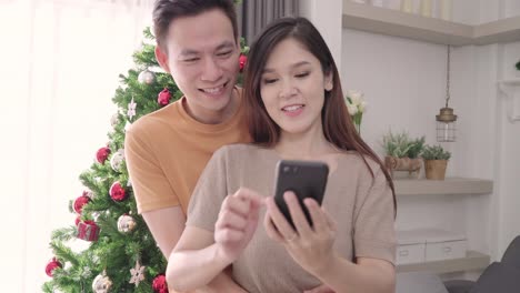 Asian-couple-using-smartphone-selfie-with-christmas-tree-decorate-their-living-room-at-home-in-Christmas-Festival.-Lifestyle-woman-and-man-happy-celebrate-summer-christmas-and-New-year-concept.