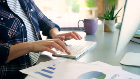 Woman-Typing-On-Computer-Next-To-Financial-Charts-At-Home-Office