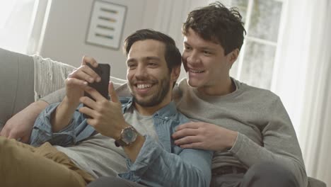 Süße-männliche-Gay-paar-verbringen-Zeit-zu-Hause.-Sie-sind-unten-auf-dem-Sofa-liegen-und-ein-Smartphone-benutzen.-Stöbern-sie-Online.-Partners-Hand-ist-um-seine-Geliebte.-Sie-Lächeln-und-lachen.-Zimmer-ist-Modern-eingerichtet.