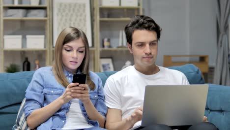 Joven-pareja-hablar-mientras-utilizando-Smartphone-y-ordenador-portátil-en-casa