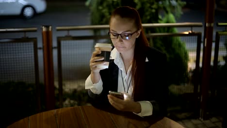 hermosa-mujer-caucásica-joven-sentada-después-del-trabajo-por-la-noche-en-un-café-en-la-calle-bebiendo-café-y-charlando-en-las-redes-sociales-usando-un-smartphone.