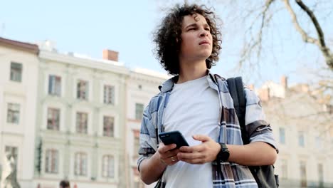 Handsome-young-man,-tourist-or-student,-millennial-in-hipster-outfit,-using-his-smartphone-scrolls-through-social-media-feed-on-device,-checking-map-or-reading-news-on-app,-looking-around