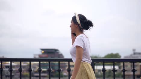 Beautiful-young-asian-woman-walking-using-smartphone-enjoying-reading-social-media-beside-fence-at-river.