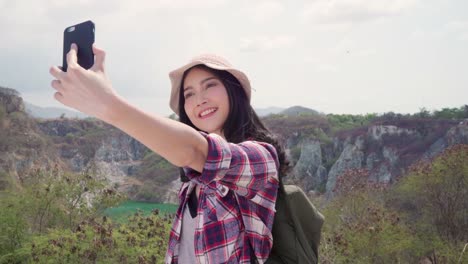 Asian-backpacker-woman-selfie-on-top-of-mountain,-young-female-happy-using-mobile-phone-taking-selfie-enjoy-holidays-on-hiking-adventure.-Lifestyle-women-travel-and-relax-concept.