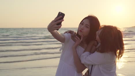 Young-Asian-lesbian-couple-using-smartphone-taking-selfie-near-beach.-Beautiful-women-lgbt-couple-happy-relax-enjoy-love-moment-when-sunset-in-evening.-Lifestyle-lesbian-couple-travel-on-beach-concept