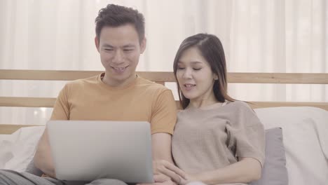 Asian-couple-using-laptop-while-lying-on-the-bed-when-relax-in-bedroom,-couple-playing-social-media-in-computer-at-home.-Couple-using-relax-time-at-home-concept.