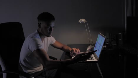 Young-afro-american-man-working-on-computer-with-bedside-lamp-at-night-in-his-dark-and-black-office.-Scrolling-on-work-documents-and-checking-messages-on-smartphone.