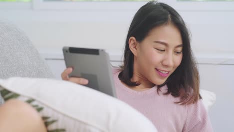 Beautiful-attractive-smiling-Asian-woman-using-tablet-holding-a-warm-cup-of-coffee-or-tea-while-lying-on-the-sofa-when-relax-in-living-room-at-home.-Lifestyle-women-at-home-concept.