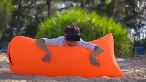 Der-junge-Mann-auf-einem-aufblasbaren-Sofa-an-einem-tropischen-Strand-verwendet-eine-VR-Brille.-Er-fühlt-sich-wie-in-einem-Meer-schwimmen-und-viele-tropische-Fische-beobachten.-Konzept-moderner-Technologien,-die-Ihnen-das-Gefühl-geben-können,-woanders-z