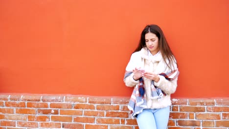 A-young-attractive-girl-stands-at-the-wall-of-a-building-and-pokes-a-finger-into-a-mobile-phone.