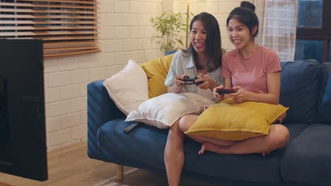 Lesbianas-mujeres-lgbt-pareja-juegan-juegos-en-casa,-mujer-asiática-usando-joystick-teniendo-divertido-momento-feliz-juntos-en-el-sofá-en-la-sala-de-estar-en-la-noche.-Joven-amante-del-fútbol-aficionado-al-fútbol,-celebrar-el-concepto-de-vacaciones.