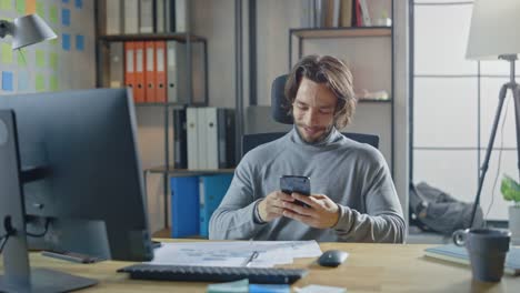 Guapo-emprendedor-de-pelo-largo-sentado-en-su-escritorio-en-la-oficina-trabaja-en-computadora-de-escritorio,-trabajando-con-documentos,-gráficos.-Sonriente-utiliza-Smartphone,-aplicación-de-medios-sociales,-escritura-de-correos-electrónicos,-mensajería