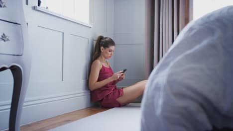 Mujer-infeliz-usando-pijama-sentado-en-el-piso-de-la-habitación-mirando-el-teléfono-móvil