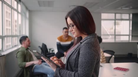 Portrait-of-young-confident-businesswoman-using-tablet-computer-smiling-and-looking-at-camera-standing-in-front-of-her-colleague-talking-in-the-background