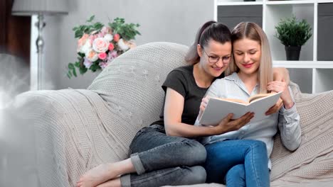 Adorable-happy-same-sex-female-couple-smiling-reading-book-together-relaxing-on-comfortable-couch