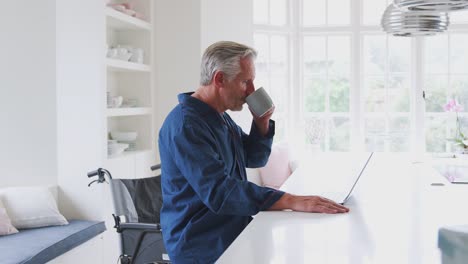 Anciano-discapacitado-hombre-en-silla-de-ruedas-en-casa-trabajando-en-el-ordenador-portátil-en-el-mostrador-de-la-cocina