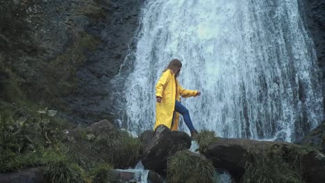 Young-girl-traveler-hiker-in-a-yellow-raincoat-walks-to-a-waterfall-in-the-highlands,-raises-his-hands-up,-enjoys-nature-and-life.-Traveling-in-the-mountains,-adventure-in-trip.-Lifestyle-concept