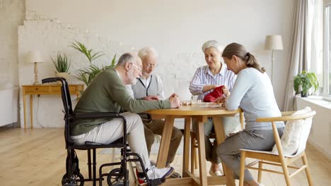 Tracking-shot-of-group-of-four-retired-elderly-people,-two-men-and-two-women,-sitting-at-table-and-playing-bingo-game-together-in-assisted-living-home