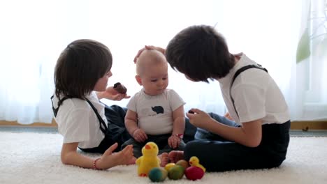 Three-cute-boys,-siblings,-brothers,-playing-with-Easter-eggs,-eating-chocolate-bunnies-at-home,-sunny-living-room