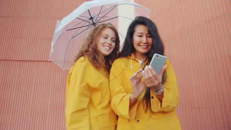 Fröhliche-junge-Frauen-in-Regenmänteln-mit-Smartphone-im-Freien-unter-Regenschirm