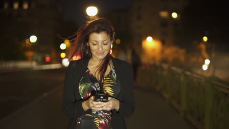 Nette-attraktive-stilvolle-kaukasische-moderne-Frau-trägt-Blumenkleid,-schwarze-Jacke-und-rote-Haare-zu-Fuß-durch-die-Straße-und-schreiben-eine-SMS-auf-ihrem-Smartphone-bei-Nacht.-Paris-4K-UHD.