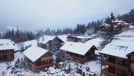 Aerial-of-wooden-cottages-in-mountain-village-surrounded-with-coniferous-forest.-Drone-view-of-chalets-covered-with-snow-at-ski-resort.-Cold-frosty-winter-day-and-snowfall-in-the-mountains