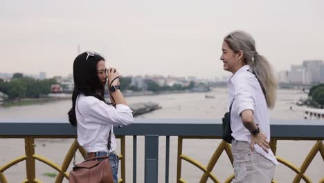 Feliz-pareja-lesbiana-asiática-tomando-una-foto-mientras-está-de-pie-en-el-puente.-Hermosas-mujeres-asiáticas-viajando-con-el-estilo-de-vida-de-vacaciones-de-un-amigo.