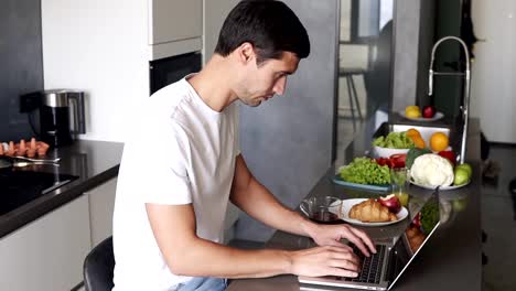 Morena-joven-sentarse-y-usar-el-trabajo-de-la-computadora-portátil-en-la-mañana-en-casa-en-el-mostrador-de-la-cocina.-Hombre-revisando-correos-electrónicos,-internet-en-las-redes-sociales,-escribiendo-mientras-bebe-un-té-de-una-taza