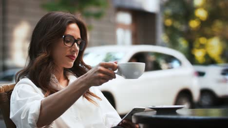 Geschäftsmädchen-in-Brille-und-weißem-Hemd.-Sie-sitzt-am-Tisch-im-Straßencafé.-Durchsuchen-von-Nachrichten-auf-dem-Handy-und-Kaffee-trinken.-Zeitlupe