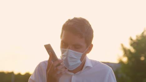 CLOSE-UP:-Mann-mit-Gesichtsmaske-ärgert-sich-beim-Telefonieren-bei-Sonnenuntergang.