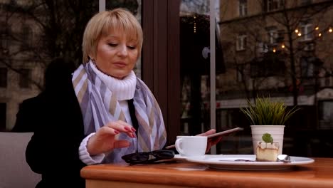 An-elderly-woman-in-a-street-cafe,-she-uses-a-tablet-and-drinks-coffee.