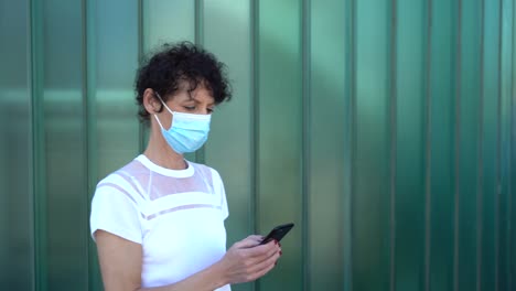 Zoom-in-shot-of-mature-woman-wearing-face-mask-while-text-messaging-on-mobile-phone-against-green-wall