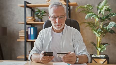 El-hombre-de-negocios-mayor-está-sosteniendo-la-tarjeta-de-crédito-e-ingresando-su-número-en-el-teléfono-inteligente,-comprando-en-línea-sentado-en-el-escritorio