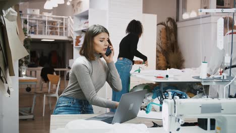 Woman-clarifying-order-details-by-smartphone-working-at-sewing-workshop,-mixed-race-seamstresses-taking-measurements-and-cutting-fabric