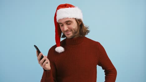 Guy-in-Santa-hat-using-smartphone,-showing-its-black-screen,-smiling-and-pointing-at-it,-raising-thumb-up,-blue-background